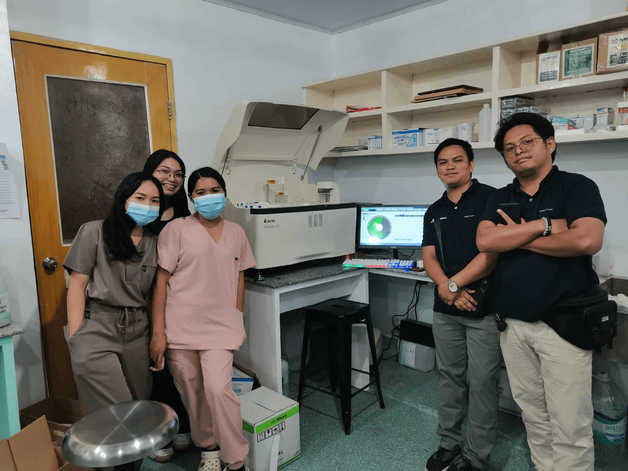 Five people beside a clinical chemistry analyzer in a laboratory.